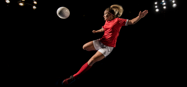 A woman wearing a red jersey kicking a soccer ball against a black background with lights shining on her.